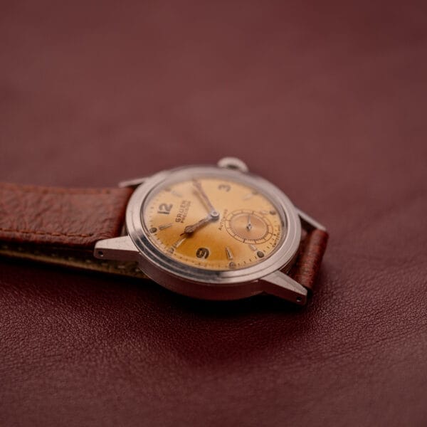 A Gruen Precision Auto-Wind Copper Dial Stainless Steel Cal. 400 wristwatch with a brown leather strap rests on a maroon leather surface.