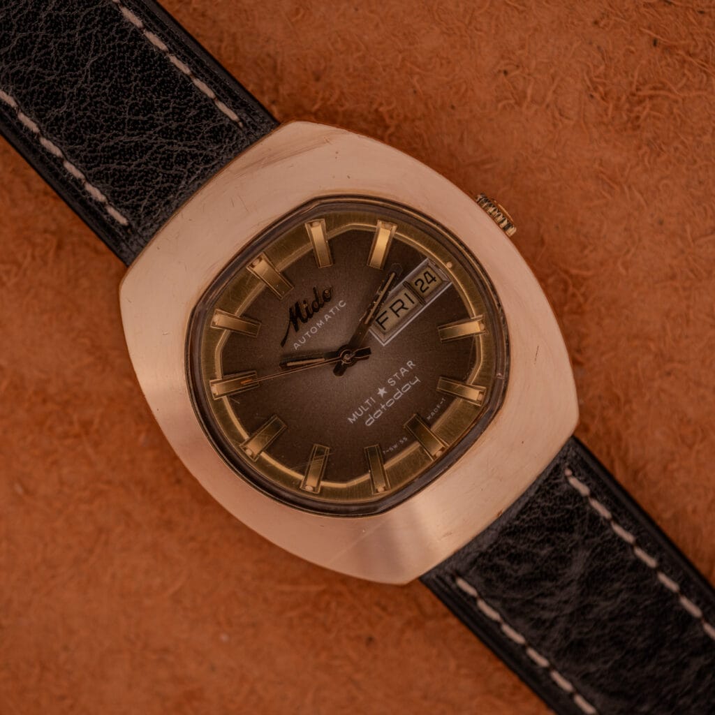 A Mido Automatic Multi Star Datoday Cal. 1819 Ref. 6929300 wristwatch with a brown face and black leather strap rests on a textured brown surface.