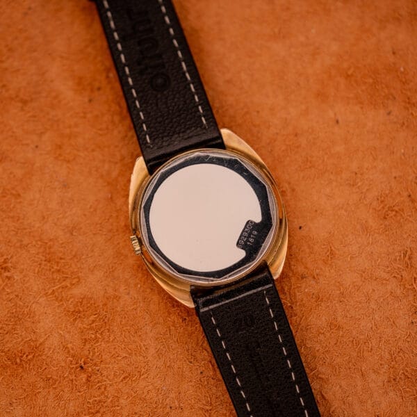 A Mido Automatic Multi Star Datoday Cal. 1819 Ref. 6929300 wristwatch with its back cover removed, revealing the intricate inner mechanism on a brown surface.