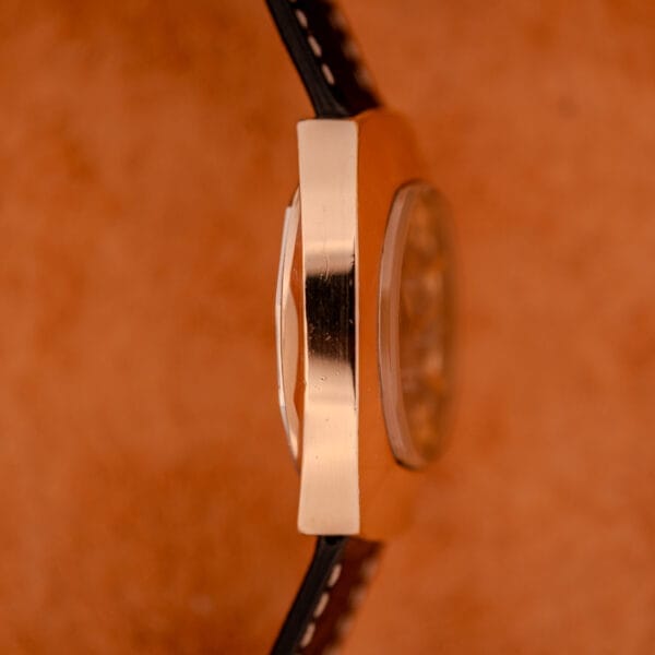 Side view of a Mido Automatic Multi Star Datoday Cal. 1819 Ref. 6929300 gold wristwatch with a black strap, set against a textured orange background.