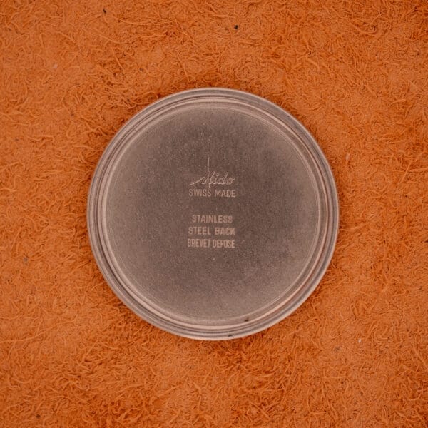 The round metal watch back of the Mido Automatic Multi Star Datoday Cal. 1819 Ref. 6929300, featuring "Mido Swiss Made" and other markings, is shown on an orange textured surface.