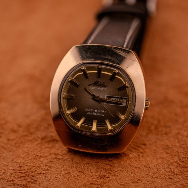 A Mido Automatic Multi Star Datoday Cal. 1819 Ref. 6929300 wristwatch with a black strap rests elegantly on a brown textured surface.