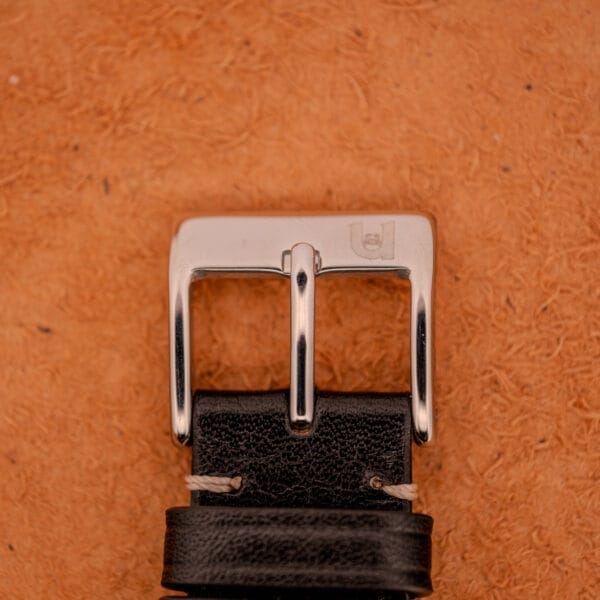 Close-up of the Mido Automatic Multi Star Datoday Cal. 1819 Ref. 6929300 silver belt buckle on a black leather strap, set against a textured brown surface.