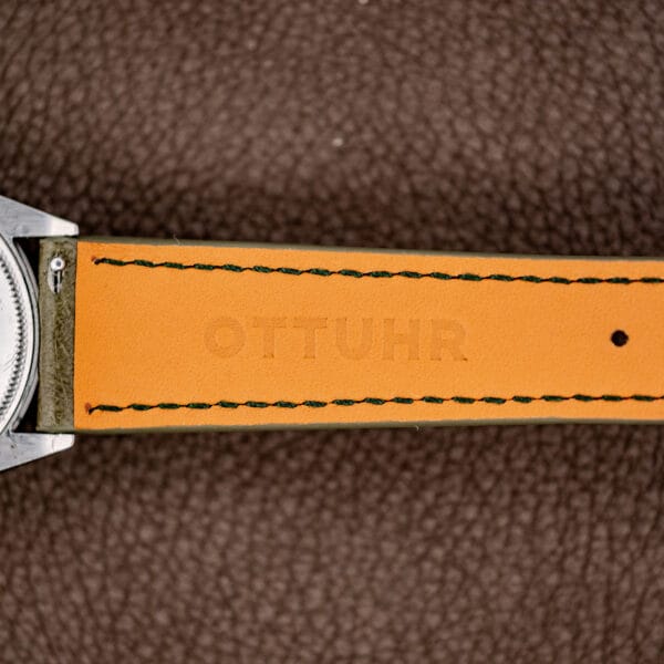 Close-up of an orange leather watch strap with OTTUHR embossed, placed on a brown textured background next to a Rolex Oyster Perpetual Air King Ref. 5500 Silver Sunburst Dial.