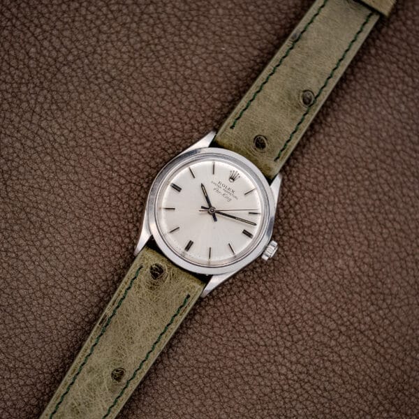 A Rolex Oyster Perpetual Air King Ref. 5500 Silver Sunburst Dial with a light green leather strap rests on a brown textured surface.