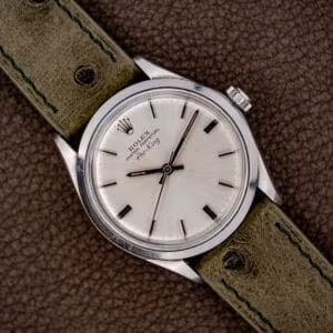 The Rolex Oyster Perpetual Air King Ref. 5500 Silver Sunburst Dial is shown with a green leather strap and displayed on brown leather for an elegant look.