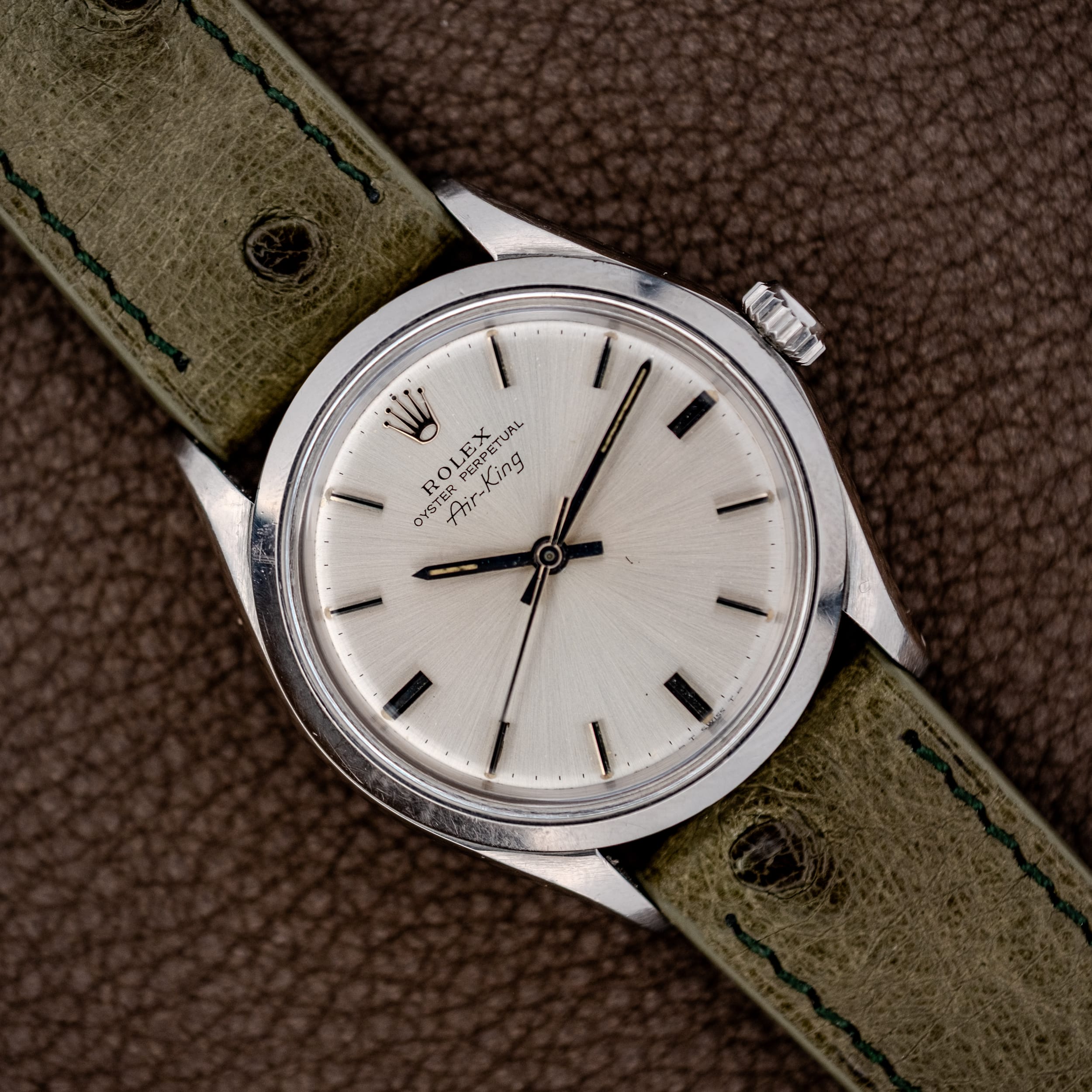 The Rolex Oyster Perpetual Air King Ref. 5500 Silver Sunburst Dial is shown with a green leather strap and displayed on brown leather for an elegant look. The Rolex Oyster Perpetual Air King Ref. 5500 Silver Sunburst Dial is shown with a green leather strap and displayed on brown leather for an elegant look.