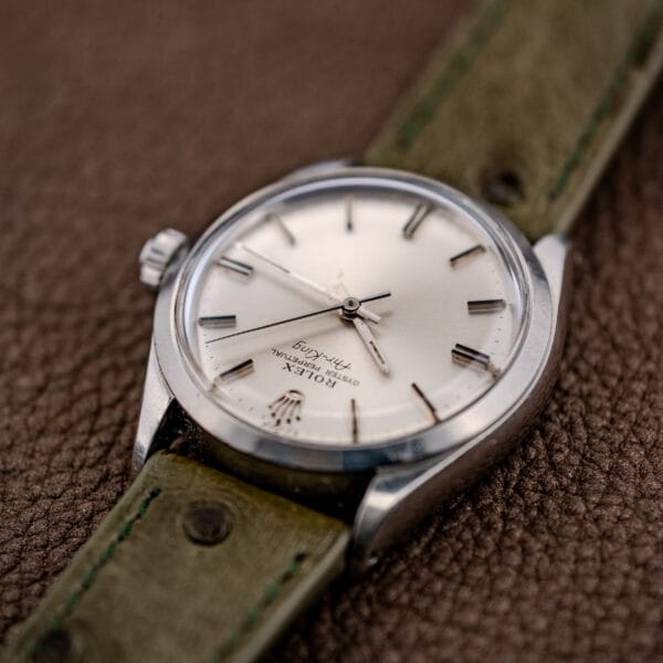 Close-up of a Rolex Oyster Perpetual Air King Ref. 5500 with a silver sunburst dial and green leather strap, displayed on a brown textured surface.