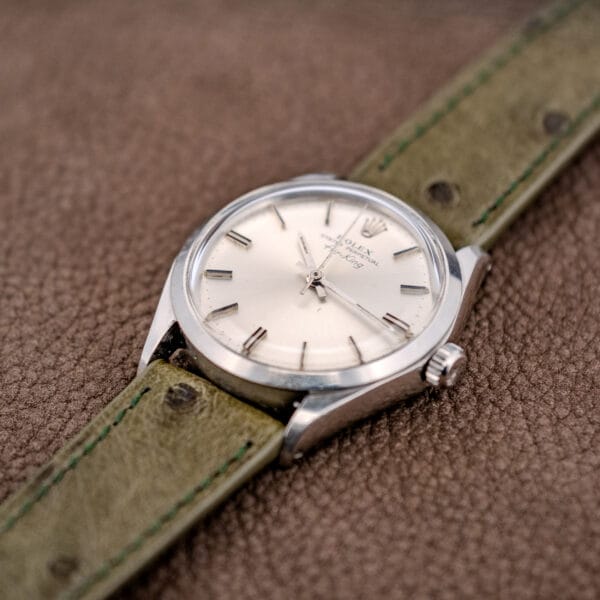 A Rolex Oyster Perpetual Air King Ref. 5500 with a Silver Sunburst Dial and light green leather strap rests elegantly on a brown textured surface.