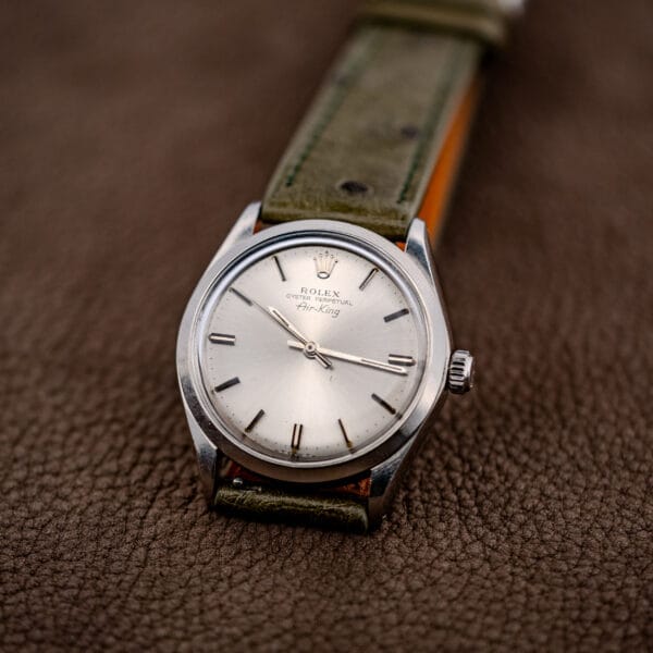 A Rolex Oyster Perpetual Air King Ref. 5500 with a Silver Sunburst Dial and green leather strap rests elegantly on a brown textured surface.