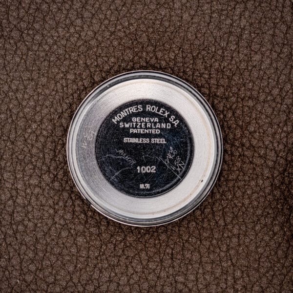 A round metal watch case back marked ROLEX GENEVA SWITZERLAND, resting on a brown textured surface, likely belongs to the Rolex Oyster Perpetual Air King Ref. 5500 Silver Sunburst Dial.