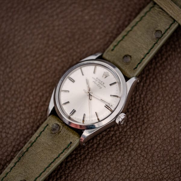A Rolex Oyster Perpetual Air King Ref. 5500 with a Silver Sunburst Dial and green leather strap is elegantly displayed on a brown textured surface.