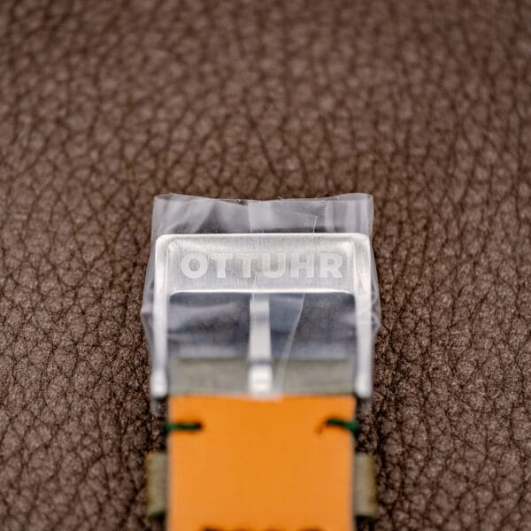 Close-up of a watch buckle labeled OTTUHR on leather, atop brown textured leather, evoking the refined details of the Rolex Oyster Perpetual Air King Ref. 5500 Silver Sunburst Dial.