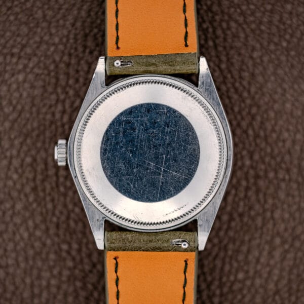 Back of a scratched Rolex Oyster Perpetual Air King Ref. 5500 Silver Sunburst Dial with an orange-brown leather strap, resting on a textured brown surface.