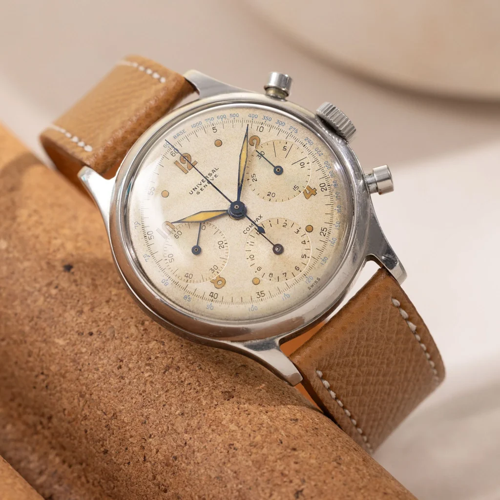 A Vintage Universal Genève stainless steel chronograph wristwatch with a tan leather strap resting on a cork surface.