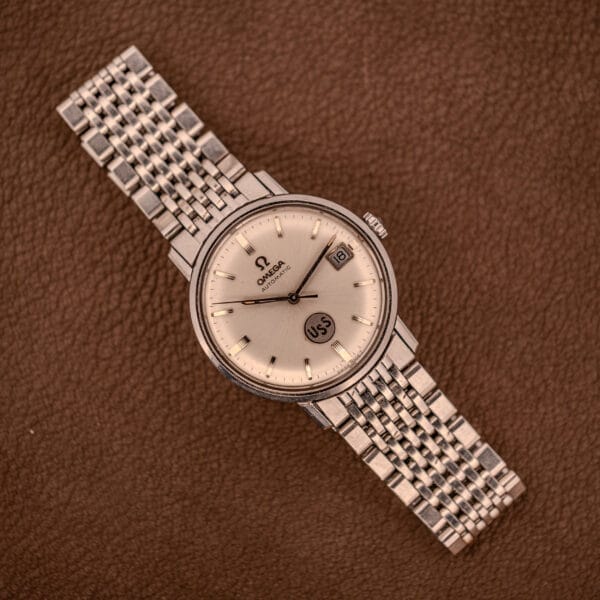 The Omega Automatic Ref. 166.020 for U.S. Steel Presentation Watch, featuring a silver case and metal band, is displayed on a brown textured surface, reflecting classic presentation watch elegance.
