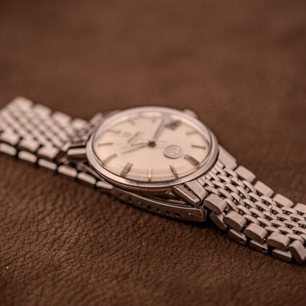 An Omega Automatic Ref. 166.020 for U.S. Steel Presentation Watch with a silver dial and metal band rests on a brown textured surface.