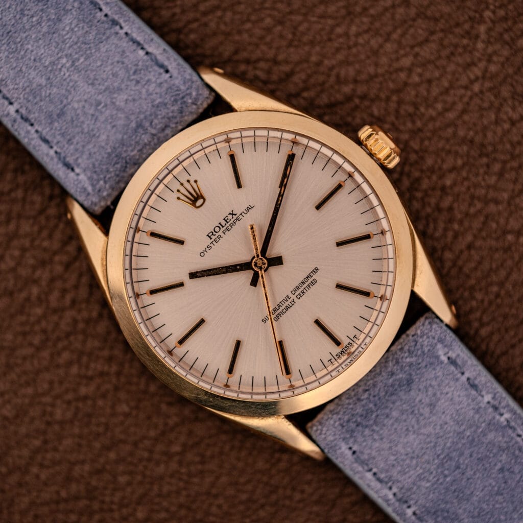 Rolex Oyster Perpetual 1024 14k Gold Capped watch with a sunburst dial and blue strap, elegantly displayed on a textured brown surface.