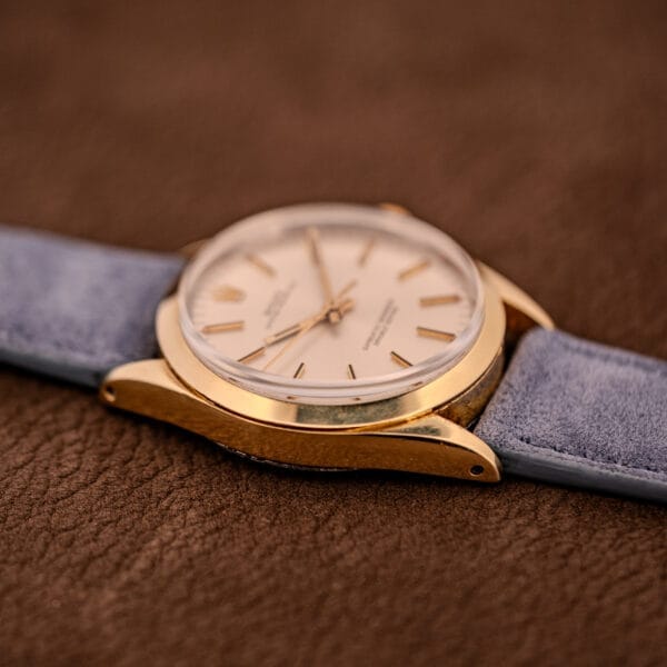 The Rolex Oyster Perpetual 1024 14k Gold Capped with a sunburst dial and light blue strap is elegantly displayed on a brown textured surface.
