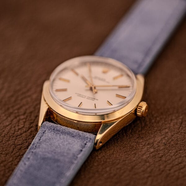 Rolex Oyster Perpetual 1024 14k Gold Capped with sunburst dial, light-colored face, and blue strap shown on a brown textured surface.