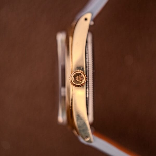 Close-up side view of a Rolex Oyster Perpetual 1024 14k Gold Capped & Sunburst Dial wristwatch crown, featuring the Rolex logo against a brown background.