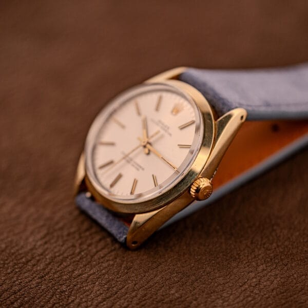 The Rolex Oyster Perpetual 1024, capped in 14k gold with a sunburst dial and blue strap, is displayed on a brown textured surface.