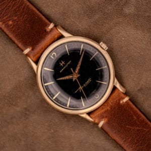 The Hamilton Thin-O-Matic Black Dial 10k Gold Filled Case Cal. 663 watch sits elegantly on a brown suede surface, featuring a sleek black dial and classic gold filled case.