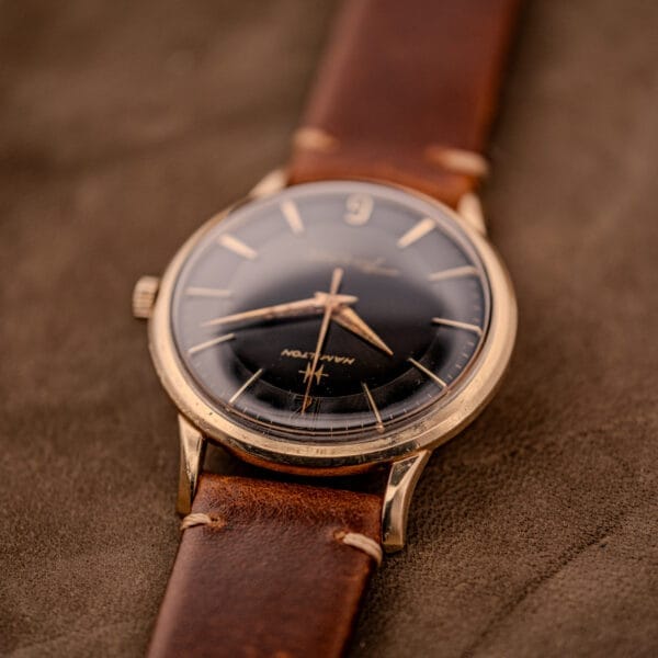 A Hamilton Thin-O-Matic Black Dial 10k Gold Filled Case Cal. 663 wristwatch with a brown leather strap is displayed elegantly on a suede surface.