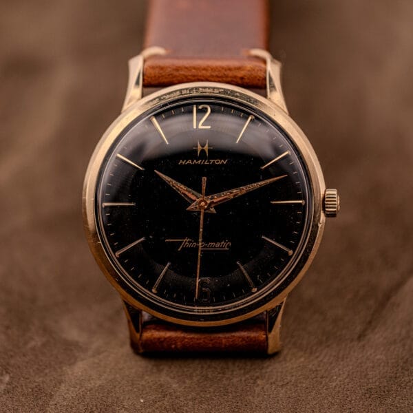 A Hamilton Thin-O-Matic Black Dial 10k Gold Filled Case Cal. 663 wristwatch with a brown leather strap is displayed on a warm brown background.