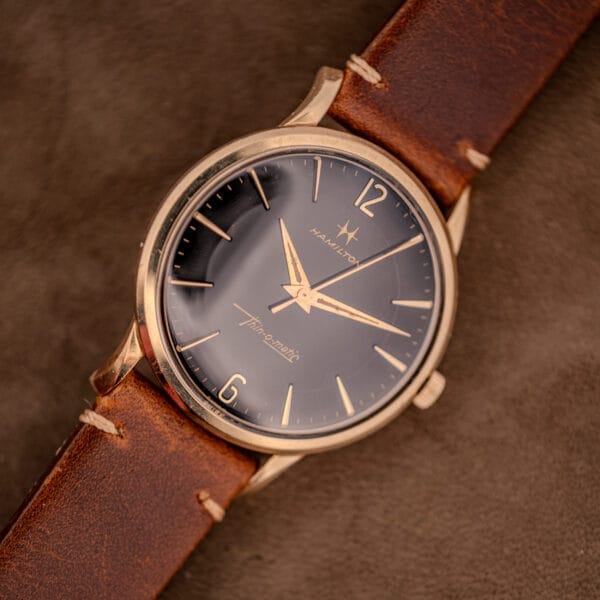 The Hamilton Thin-O-Matic Black Dial 10k Gold Filled Case Cal. 663 wristwatch features a striking black dial, brown leather strap, and elegant gold-filled case, beautifully displayed on a brown surface.