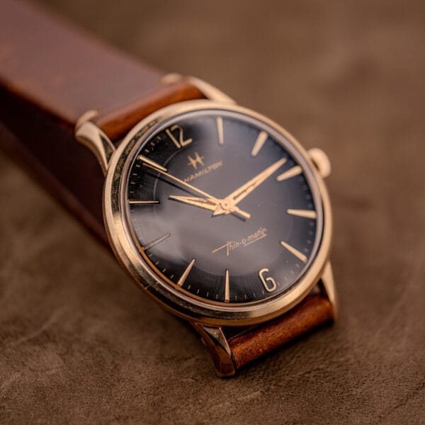 Hamilton Thin-O-Matic Black Dial 10k Gold Filled Case Cal. 663 wristwatch with a brown leather strap, displayed on a brown surface.