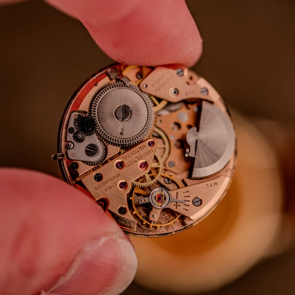 A close-up of fingers holding an intricate Hamilton Thin-O-Matic Black Dial 10k Gold Filled Case Cal. 663 watch movement with visible gears and components.
