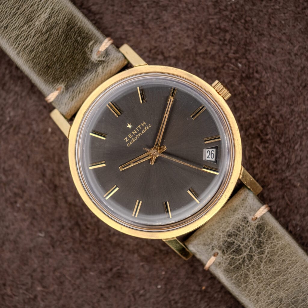 The Zenith Automatic Date Charcoal Sunburst Dial Cal. 2542 PC is a vintage dress watch with a gold case, charcoal sunburst dial, date window, and Cal. 2542 PC movement, paired with an olive green leather strap.
