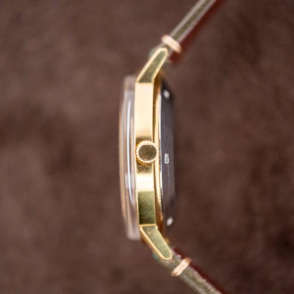 Side view of a Zenith Automatic Date watch with a charcoal sunburst dial and Cal. 2542 PC movement, featuring a gold case and brown leather strap against a blurred brown background.