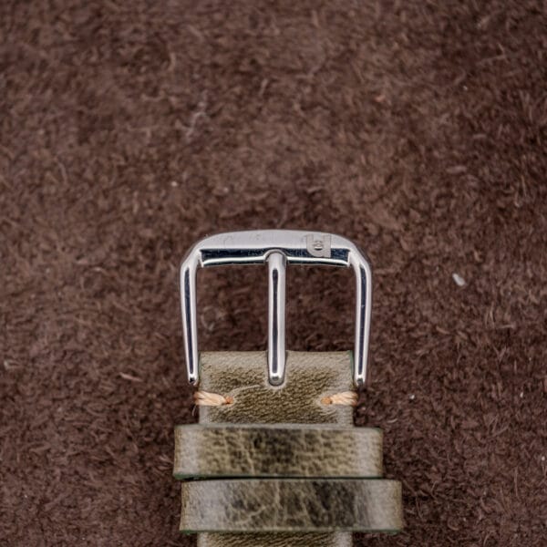 Close-up of a silver belt buckle on a green leather belt atop a brown textured surface—its polished shine echoing the charcoal sunburst dial of the Zenith Automatic Date Cal. 2542 PC.