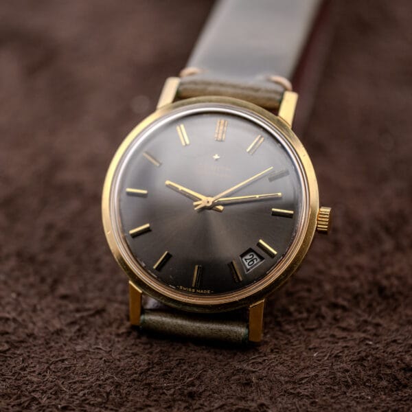 A Zenith Automatic Date Charcoal Sunburst Dial Cal. 2542 PC gold wristwatch is shown resting on a brown textured surface.