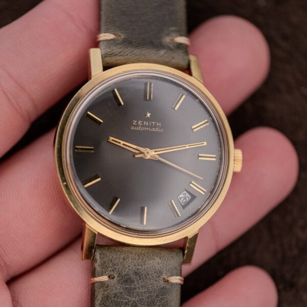 A hand holding a Zenith Automatic Date Charcoal Sunburst Dial Cal. 2542 PC wristwatch with a gold case and leather strap.