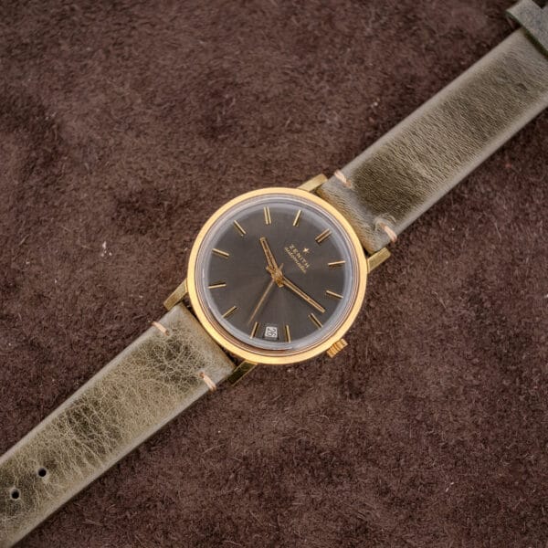 A Zenith Automatic Date Charcoal Sunburst Dial Cal. 2542 PC wristwatch with a green leather strap rests on a textured brown surface.