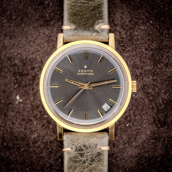 A Zenith Automatic Date Charcoal Sunburst Dial Cal. 2542 PC vintage wristwatch featuring a gold case, green leather band, and date display at 4 o’clock.
