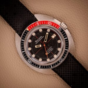 A vintage Bulova Oceanographer Automatic Snorkel 714 Red Cross Hair Dial watch featuring a black dial, red and black bezel, and a textured black strap.