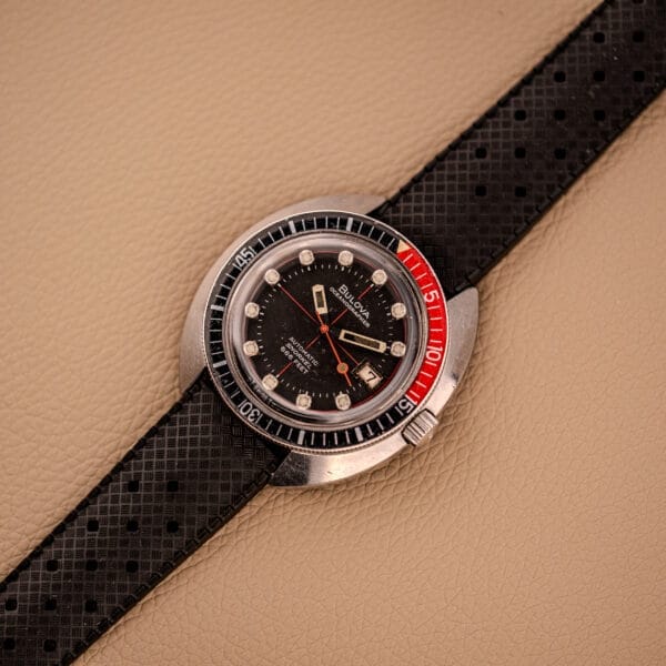 A Bulova Oceanographer Automatic Snorkel 714 with a Red Cross Hair Dial, black strap, and red-black bezel rests elegantly on a textured beige surface.
