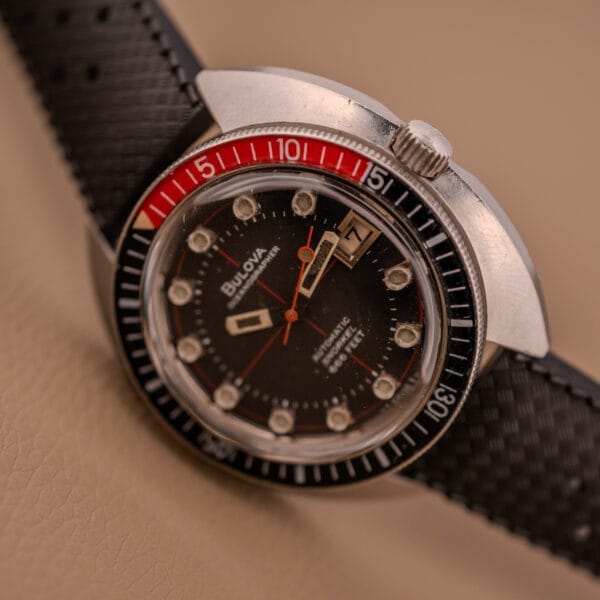 A close-up of the Bulova Oceanographer Automatic Snorkel 714 with a red cross hair dial, black and red bezel, and textured black strap.