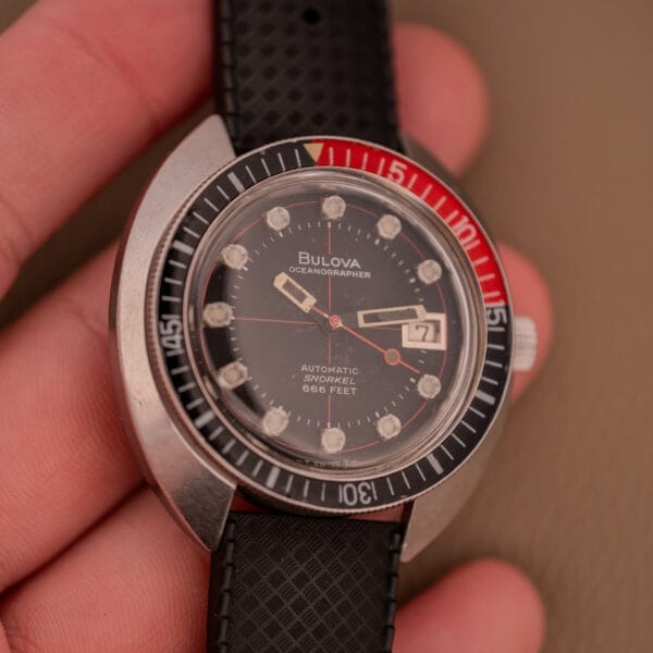 Hand holding a Bulova Oceanographer Automatic Snorkel 714 Red Cross Hair Dial watch with black/red bezel and textured black strap.
