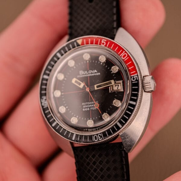 A Bulova Oceanographer Automatic Snorkel 714 wristwatch with a red cross hair dial is held in a person's hand.