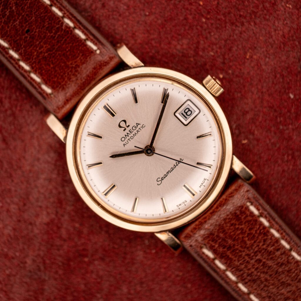 An Omega Automatic Seamster Date Ref. KM 6323 Cal. 563 with a brown leather strap rests on a red textured surface, showing the date as the 18th.