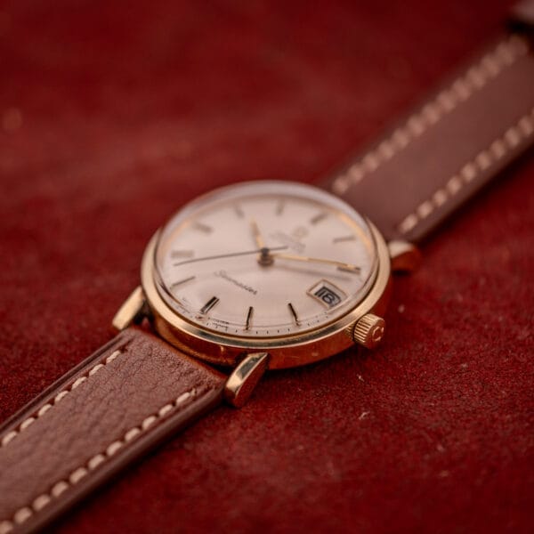 The Omega Automatic Seamaster Date Ref. KM 6323 Cal. 563 gold wristwatch with a brown leather strap is shown on a red textured surface.