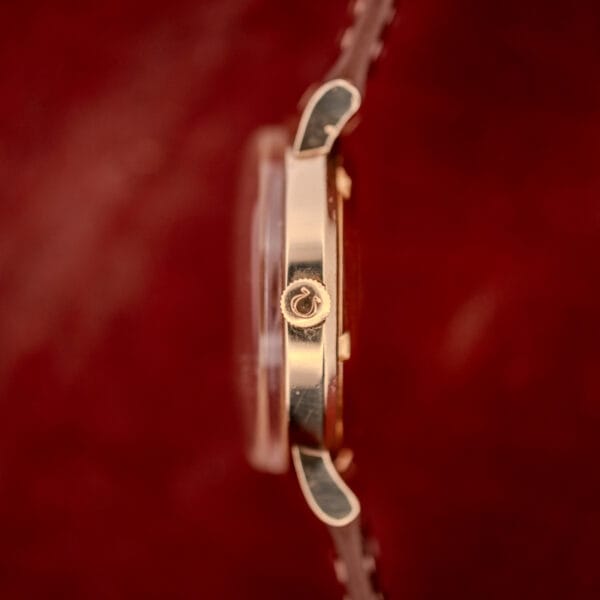 Side view of an Omega Automatic Seamster Date Ref. KM 6323 Cal. 563 gold watch, featuring an engraved crown symbol, photographed against a red background.