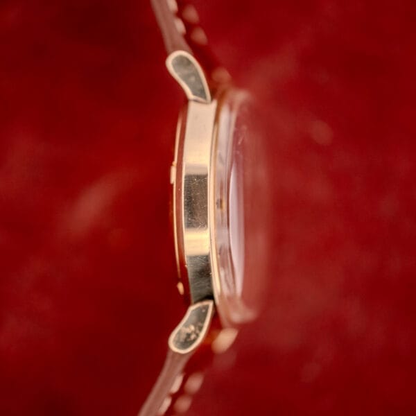 Close-up side view of the Omega Automatic Seamaster Date Ref. KM 6323 with gold case and lugs, showcasing the Cal. 563 automatic movement against a vivid red background.