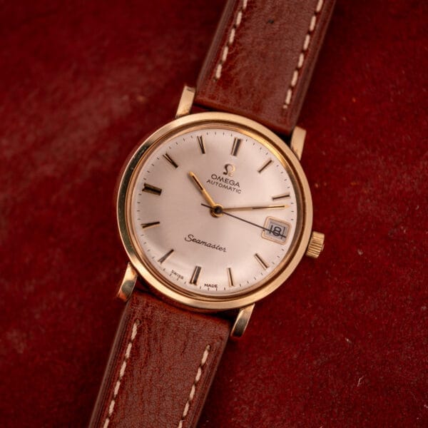 Omega Automatic Seamaster Date Ref. KM 6323 with Cal. 563 movement and a brown leather strap, displayed on a red textured background.