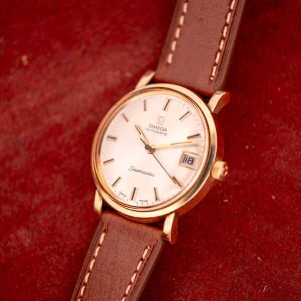 An Omega Automatic Seamaster Date Ref. KM 6323 with a Cal. 563 movement and brown leather strap rests on a red textured surface.